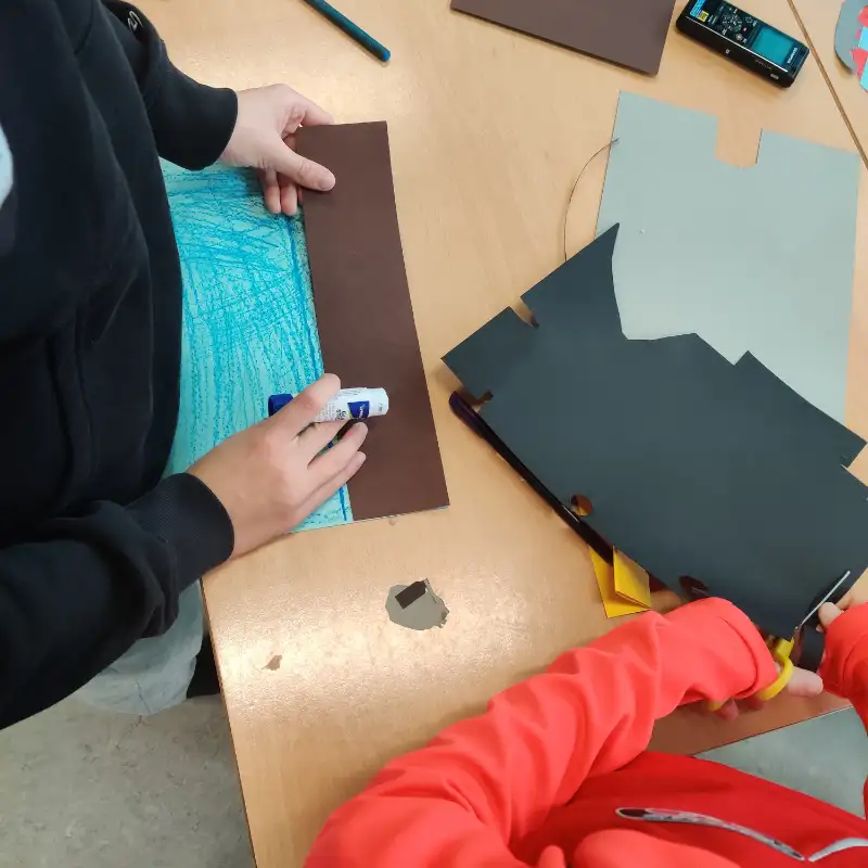 Elever arbejder med håndværk på et bord, klipper og limer papir i et skoleklasseværelse.